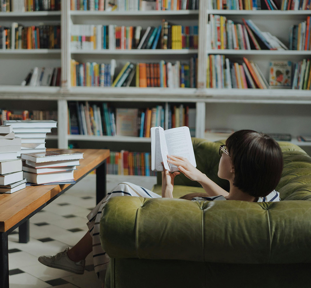 A person sitting comfortably on a green couch reading a book. In the background are bookshelves full of more reading options.