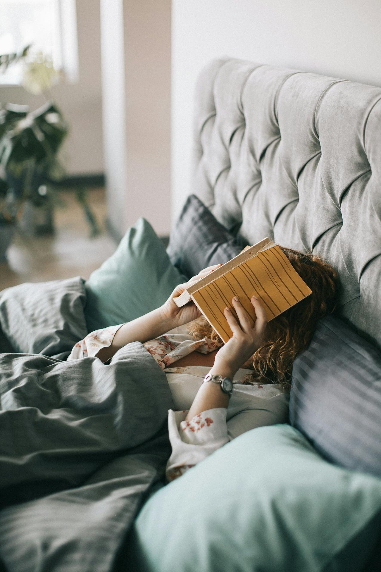 An image showing social burnout with a woman in bed and a book over her face. Ready to relax and recuperate for her mental health